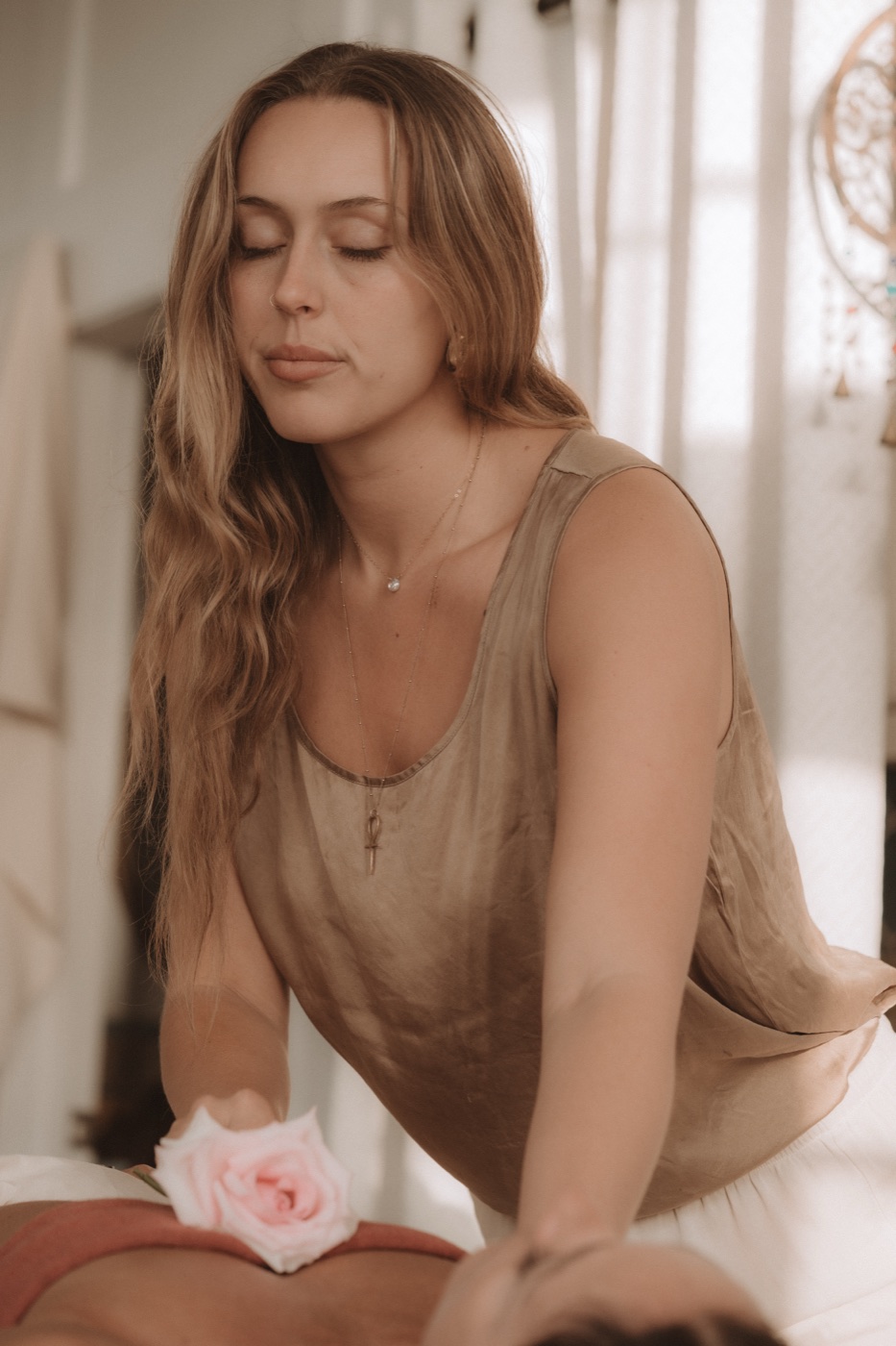 Alaya at the table, eyes closed, hands resting at a client's back with a pale rose laid on the drape