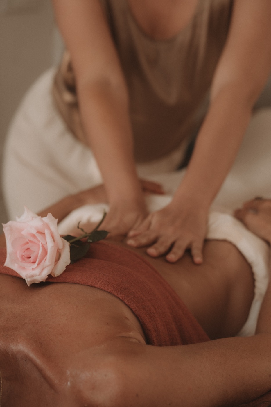 Alaya's hands working slowly across a draped back, a pale pink rose resting on the linen