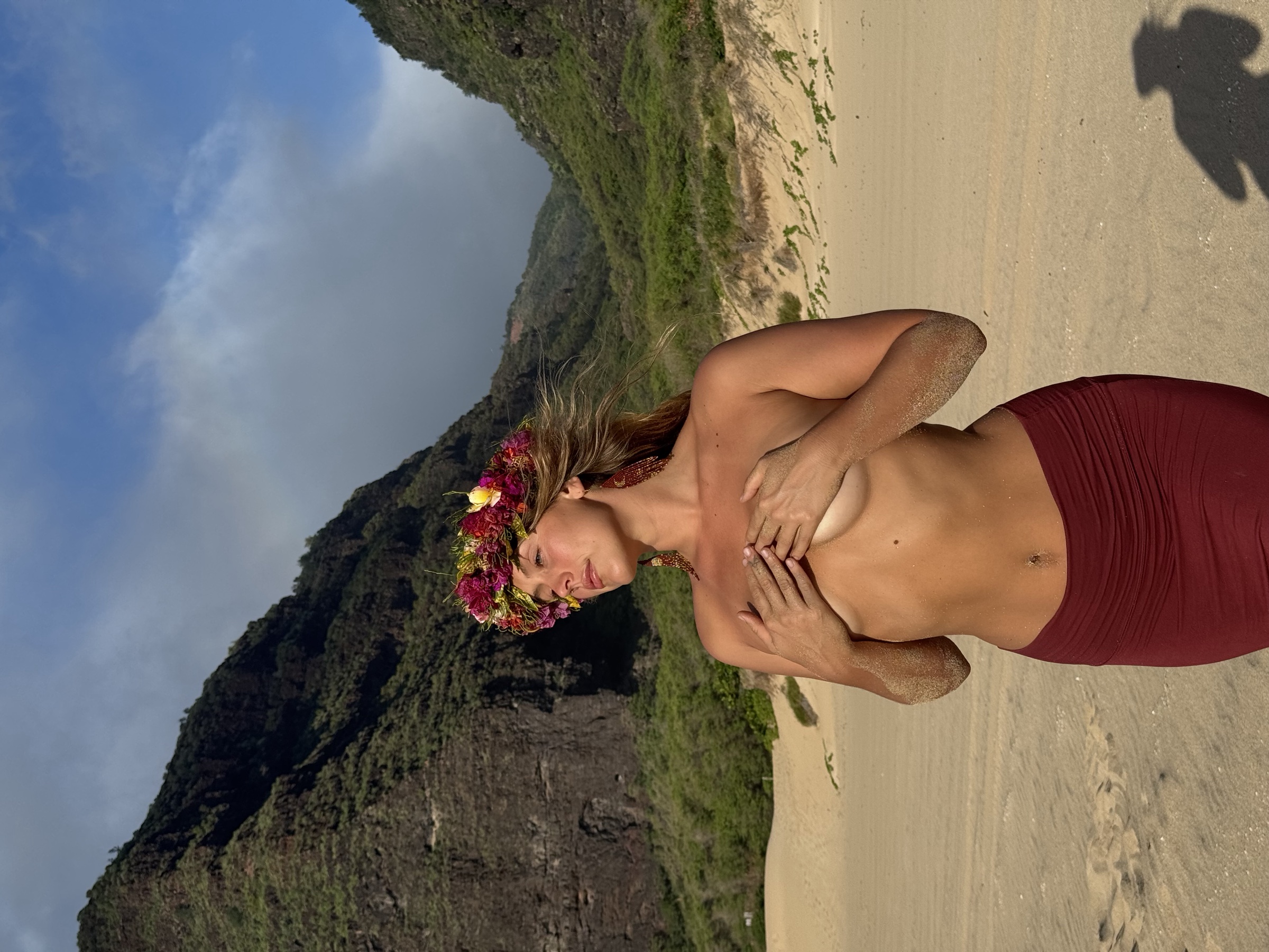 Alaya in flower crown, hands at the heart, sand and mountain coastline behind her